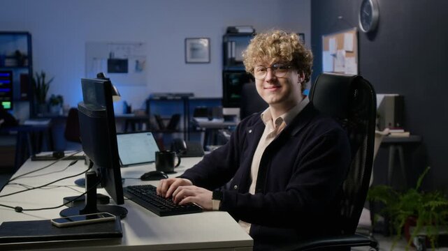 Portrait of young adult Caucasian IT analyst typing on keyboard and turning to smile at camera in office. Perfect for cybersecurity recruiting and corporate technology marketing campaigns