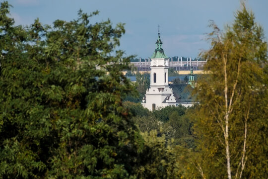 Kolegiata św. Michała Archanioła w Ostrowcu Świętokrzyskim p&oacute;źnym latem. Obiekt wyłania się spośr&oacute;d bujnej zieleni; wyr&oacute;żnia się tradycyjną architekturą, wysoką białą wieżą oraz  miedzianym dachem