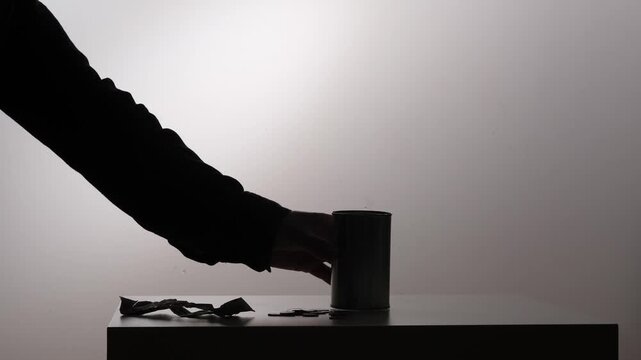 Hand pouring the last few coins from a small tin can onto a surface, highlighting concepts of financial hardship, dwindling savings, and economic struggle against a backlit silhouette