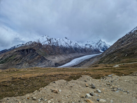 View of the Penzi La road winding through the rugged mountain landscape of Zanskar, featuring snow-capped peaks and a glacier under a cloudy sky in Zanskar, Ladakh, India.