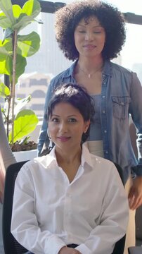 Vertical video: Smiling woman in denim urging peer in white top posing and laughing at office chair