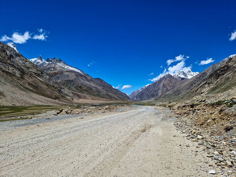View of the Penzi La road winding through a high-altitude mountain valley with snow-capped peaks and rocky terrain under a clear blue sky in Zanskar, Ladakh, India.