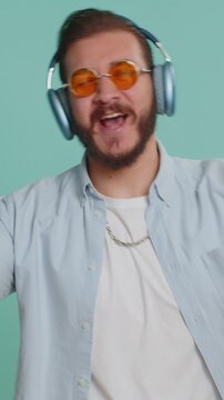 Happy Lebanese man in headphones listening music and dancing disco fooling around having fun expressive gesticulating hands relaxing on party. Arabian boy isolated alone on studio blue wall background