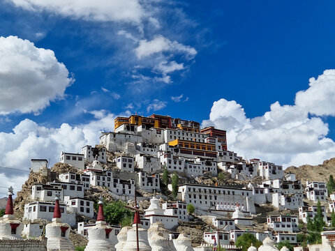 View of Thiksey Monastery, a large Tibetan Buddhist monastery complex with white-washed buildings and red roofs, set against a bright blue sky in Thiksey, Ladakh, India.