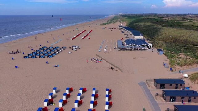 High-angle aerial drone view of Katwijk aan Zee beach resort in South Holland Netherlands with beach pavilion, red windbreak cabins, blue sun loungers, dunes, and North Sea in summer sunlight