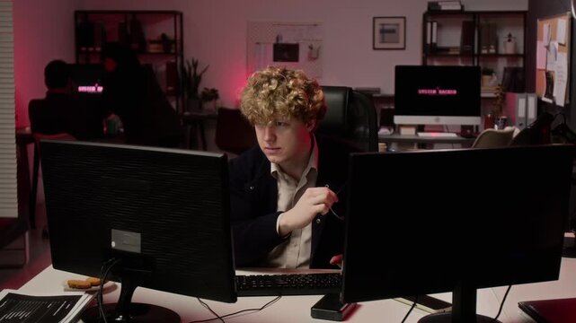 Caucasian young adult cybersecurity analyst taking off glasses while looking at computer screen during cyber attack. Footage suitable for data breach, IT security and network defense campaigns