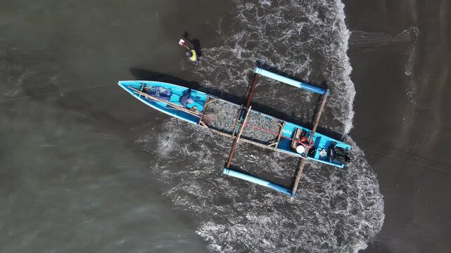Aerial view of two blue outrigger fishing boats resting on the dark sand beach as waves wash ashore in Cilacap, Central Java, Indonesia.