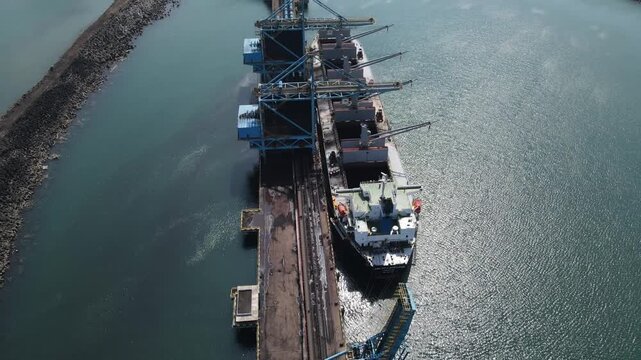Aerial view of a large cargo ship docked at an industrial port terminal with loading cranes and calm water in Cilacap, Central Java, Indonesia.