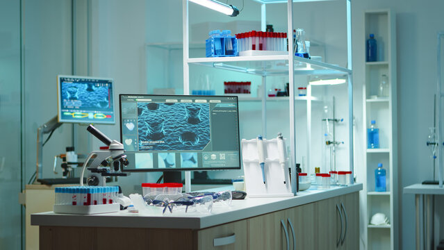 Wide empty laboratory interior with microscope, sample racks, pipettes and computer screens arranged around a modern biotechnology research bench.