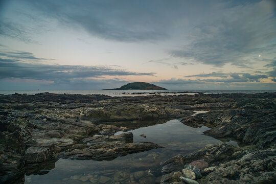 View of Looe Island, also known as St Georges Island, seen from the rocky shoreline of Hannafore beach at dusk with calm tide pools and a soft twilight sky in Looe, England, United Kingdom.