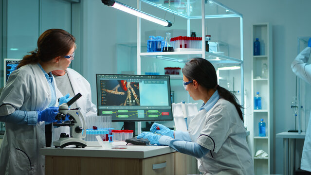 Two female scientists coordinate microscope work and computer data review beside specimen tubes during collaborative biomedical research.