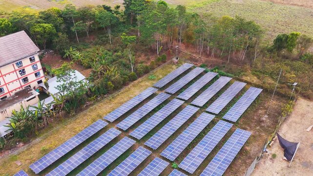 Aerial view of a solar panel farm with rows of photovoltaic modules situated near a building and lush trees in Malang, East Java, Indonesia.