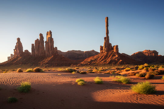 View of the Totem Pole sandstone spire in Monument Valley, surrounded by desert landscape with red sand, sparse green shrubs, and clear blue sky in Navajo County, Arizona, United States.
