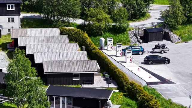 Aerial view of a row of small wooden cabins next to an electric vehicle charging station with parked cars in Norway.