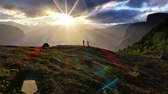 Aerial view of two people walking along a grassy mountain ridge during a vibrant sunset with lens flare in Aurland, Vestland, Norway.