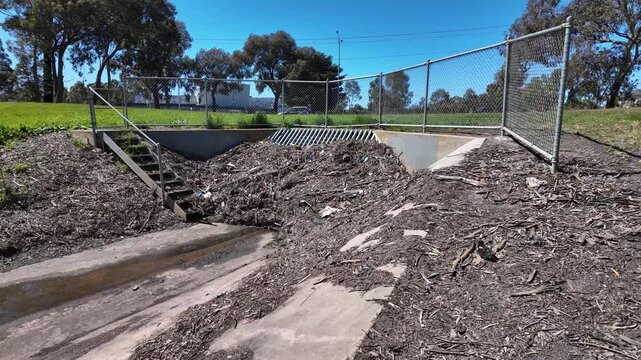 A suburban stormwater drain and culvert outlet in Melbourne, Australia, clogged with a pile of debris, leaves, and litter against a grate. Blocked drainage and flash flooding hazards