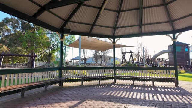 Interior view looking out from inside a large wooden gazebo shelter, featuring built-in perimeter wooden seating,&nbsp;overlooking a children&rsquo;s playground at a town park in Keith, South Australia