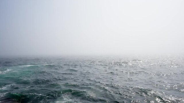 Dense sea fog rolling over dark, choppy ocean waters with sunlight glistening on the rolling waves.