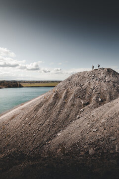 View of Aidu, a large rocky hill with two people standing on the summit, overlooking a blue lake and a dirt path in Aidu, Ida-Viru County, Estonia.