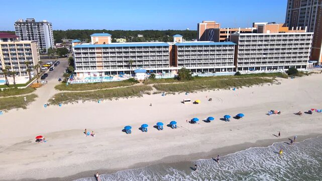 Blue umbrellas line the sand below long hotel balconies during a lateral aerial pass.
