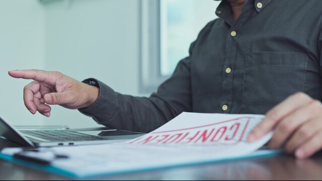 Businessman reviewing confidential document paper on clipboard beside laptop at corporate office desk, pointing finger forward during official private audit assessment procedure.