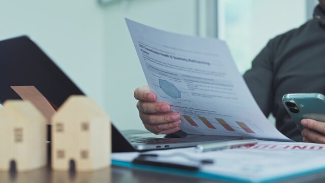 Real estate investor reviewing financial market reports beside miniature wooden houses in office setting.