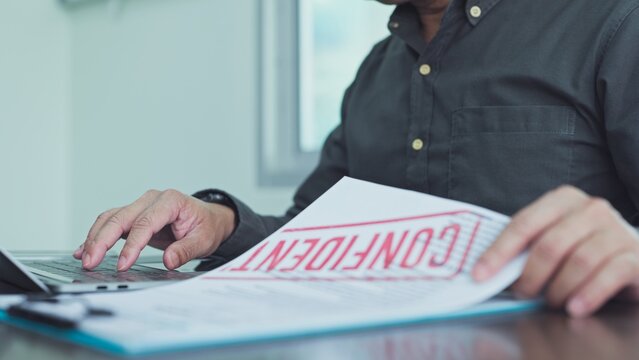 Businessman working on laptop computer reviews confidential document stamped in red ink at desk in workplace office setting, handling private corporate records carefully.