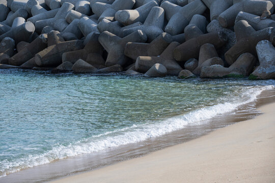 Concrete tetrapod breakwater protecting coastal shoreline with clear blue ocean waves washing over sandy beach in sunlight.