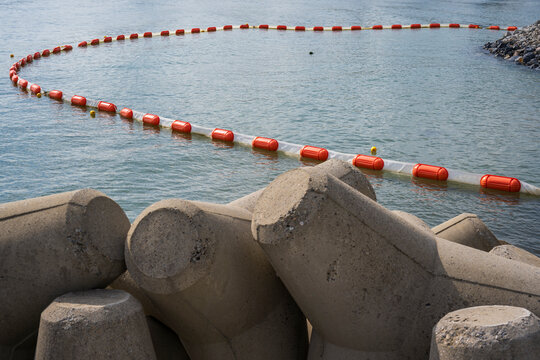 Concrete tetrapod breakwater with orange floating oil containment boom barrier on calm sea water surface along coastal harbor.