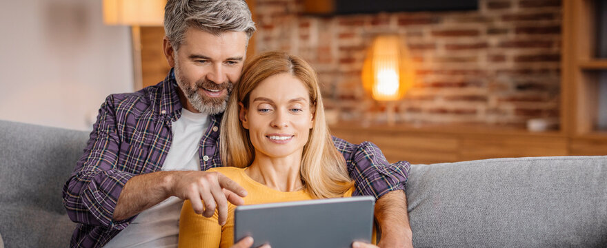 Happy Caucasian Couple, Middle Aged, Smiling, Spending Quality Time Together While Using Tablet In Cozy Living Room Setting. Enjoying Relaxed Atmosphere, Sharing Content, Technology Concept.