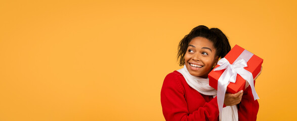 Fototapeta premium Smiling Young Black Woman Expressing Joy While Holding Red Gift Box With White Ribbon, Standing Against Bright Yellow Background, Signifying Celebration And Festivity, Gift Giving Concept.