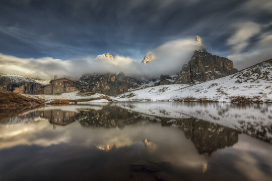View of Baita Segantini reflected in a calm alpine lake with snow-dusted mountains and dramatic clouds in Passo Rolle, Trentino-Alto Adige, Italy.