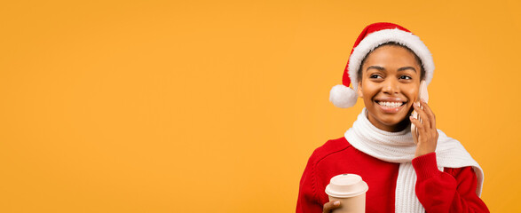 Fototapeta premium Smiling Young African American Woman Wearing Santa Hat While Talking On Phone With Coffee Cup In Hand, Standing Against Yellow Background, Embracing Holiday Spirit In A Festive Setting.