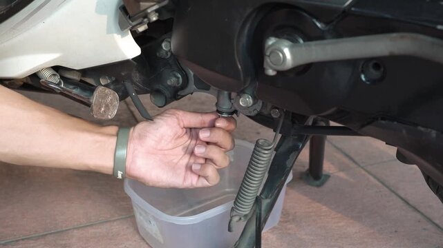 A mechanic's hand loosens the oil drain plug during a motorcycle engine oil change in a home garage.