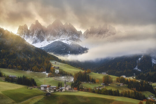 View of the Church of St. Magdalena nestled in the green meadows of the Dolomites with snow-capped peaks and dramatic clouds in the background in St. Magdalena, Trentino-Alto Adige, Italy.