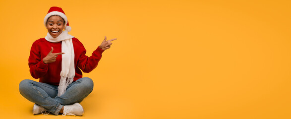Fototapeta premium Smiling Black Woman Wearing Santa Hat Sitting Cross-legged On Studio Floor, Pointing Gesture With Hands In Festive Yellow Background, Expressing Cheerfulness And Joy For Christmas Celebration.