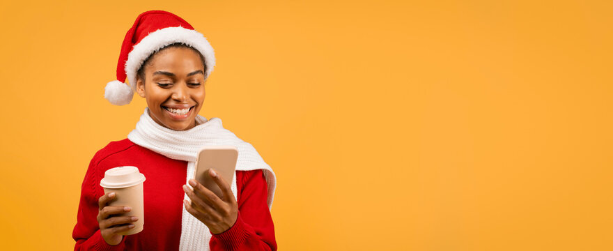Smiling African American Woman In Santa Hat Holding Coffee Cup And Looking At Smartphone In Holiday Setting. Joyful Female Enjoying Communication At Home, Celebrating Festive Season.
