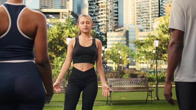 Instructor balances in tree pose with participants following the same position on yoga mat during fitness class outdoors in public park, supporting a balance pose with hands held in prayer.