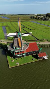 Vertical aerial drone view of De Kat historic paint windmill in Zaanse Schans Zaandam North Holland Netherlands with thatched roof orange sails green polder fields and canals on sunny day