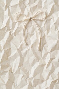 Crumpled cream paper texture with white string bow. Tied twine on wrinkled off white background. Minimalist rustic surface with natural folds, shadows. Blank organic paper material for craft design,