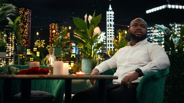 Lonely man waits alone at a rooftop restaurant table after being stood up for a date and feels distressed when his girlfriend misses their date night.