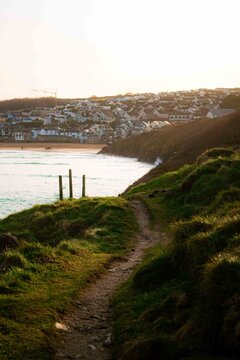 View of a coastal path leading towards a sandy beach and a town nestled on the hillside under warm golden hour light, United Kingdom.