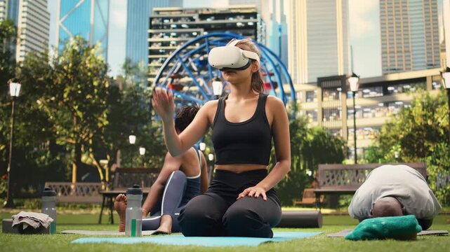 Woman wearing a virtual reality headset reaches forward with one hand while kneeling on yoga mat during immersive outdoor fitness session in city park, supporting workout simulation in VR.