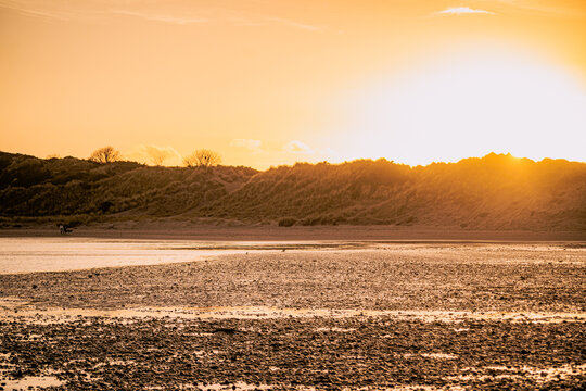 View of a sandy beach with pebbles at low tide, backed by grassy dunes under a warm golden sunset, United Kingdom.