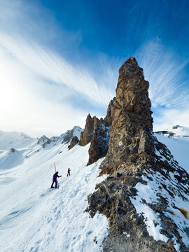 View of a rocky mountain peak covered in snow with two skiers traversing the slope under a bright blue sky in the United Kingdom.