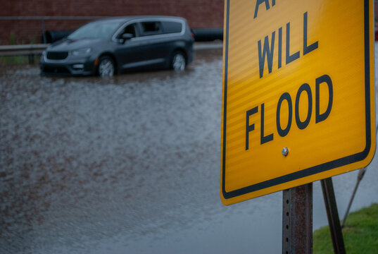 Yellow Flood Warning Sign Foregrounded with a Stranded Minivan Submerged in Deep Water on a Flooded Street