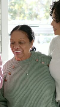 Vertical video: Smiling elder turning near window in green, pendant seen as adult leaning in white