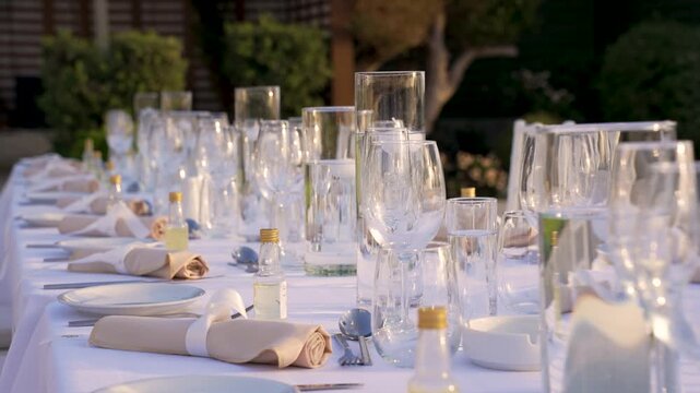 Tables are arranged in a garden for a gathering. Clear glasses and plates are set at each place with napkins and bottles prepared for guests.