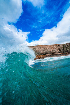 View of a powerful turquoise wave breaking near the rocky coastline under a bright blue sky with fluffy white clouds in Newquay, England, United Kingdom.