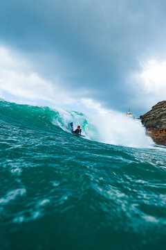 View of a bodyboarder riding a powerful turquoise wave with rocky cliffs in the background under a cloudy sky in Newquay, England, United Kingdom.